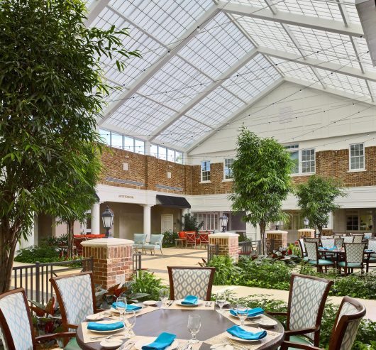 Interior view of Still Hopes Retirement Community featuring a translucent skyroof, lush greenery, and dining area with blue accents, emphasizing natural daylighting design by Kalwall.