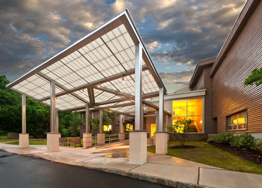 Temple Shalom exterior featuring Kalwall canopy set against green trees, cloudy sky, and lit building interior