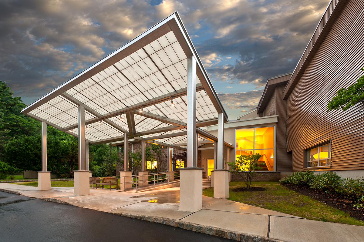 Temple Shalom exterior featuring Kalwall canopy set against green trees, cloudy sky, and lit building interior