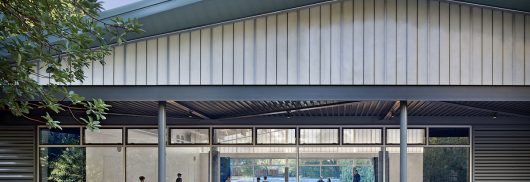 Community center exterior featuring tree and Kalwall facade w/ view to interior of building with people moving around inside.