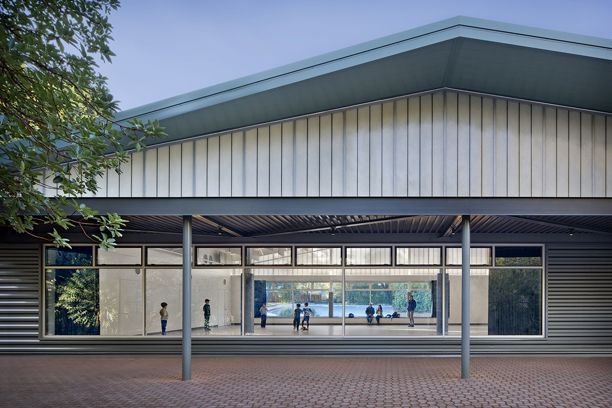 Community center exterior featuring tree and Kalwall facade w/ view to interior of building with people moving around inside.