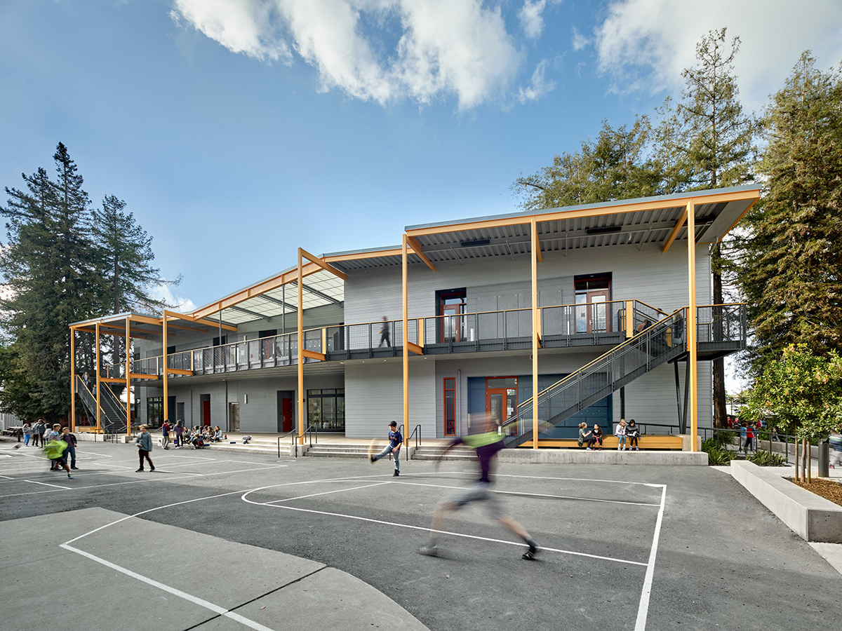 Exterior of school playground yard with concrete court with kids playing and gray building in background with Kalwall canopy.