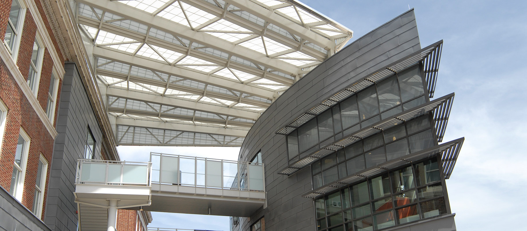 Exterior view of a brick building connected to a dark gray building by a walkway and a Kalwall canopy