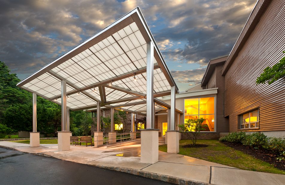 Temple Shalom exterior featuring Kalwall canopy set against green trees, cloudy sky, and lit building interior