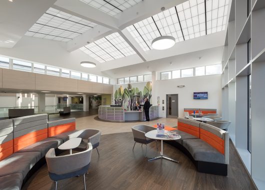 Bright, modern healthcare facility interior featuring ample natural light from clerestory windows and skylights, promoting visual comfort and wellness, with seating areas and a reception desk.
