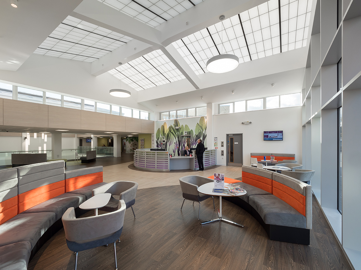Bright, modern healthcare facility interior featuring ample natural light from clerestory windows and skylights, promoting visual comfort and wellness, with seating areas and a reception desk.