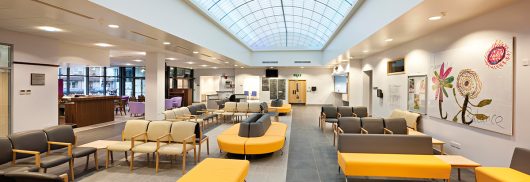 Interior view of Loxford Polyclinic showcasing modern seating arrangements with gray and yellow furniture, translucent ceiling panels for natural light, and an information point, emphasizing Kalwall's innovative facade solutions.