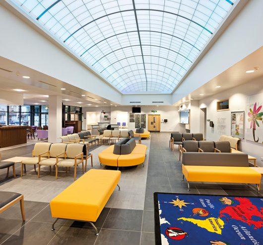 Interior view of Loxford Polyclinic showcasing modern seating arrangements with gray and yellow furniture, translucent ceiling panels for natural light, and an information point, emphasizing Kalwall's innovative facade solutions.