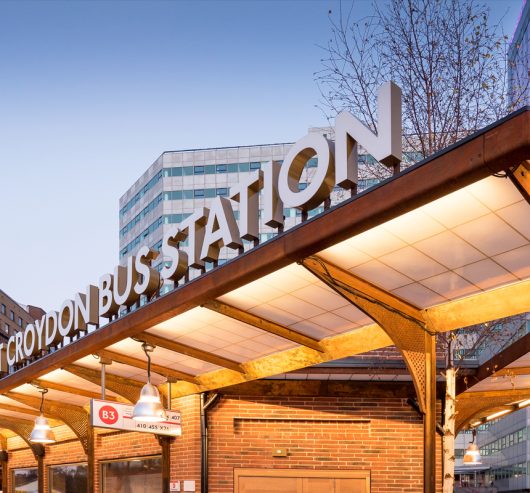 Nighttime view of West Croydon Bus Station sign atop a Kalwall canopy for a brick building, with a cityscape background