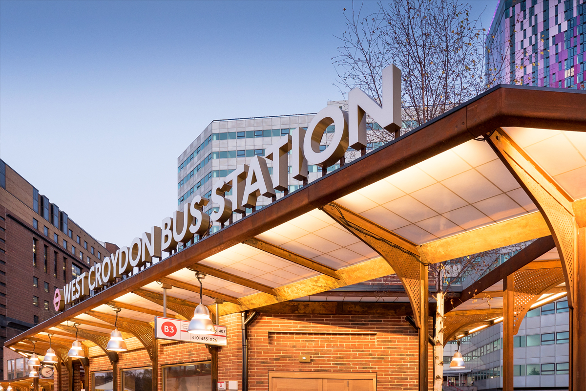 Nighttime view of West Croydon Bus Station sign atop a Kalwall canopy for a brick building, with a cityscape background