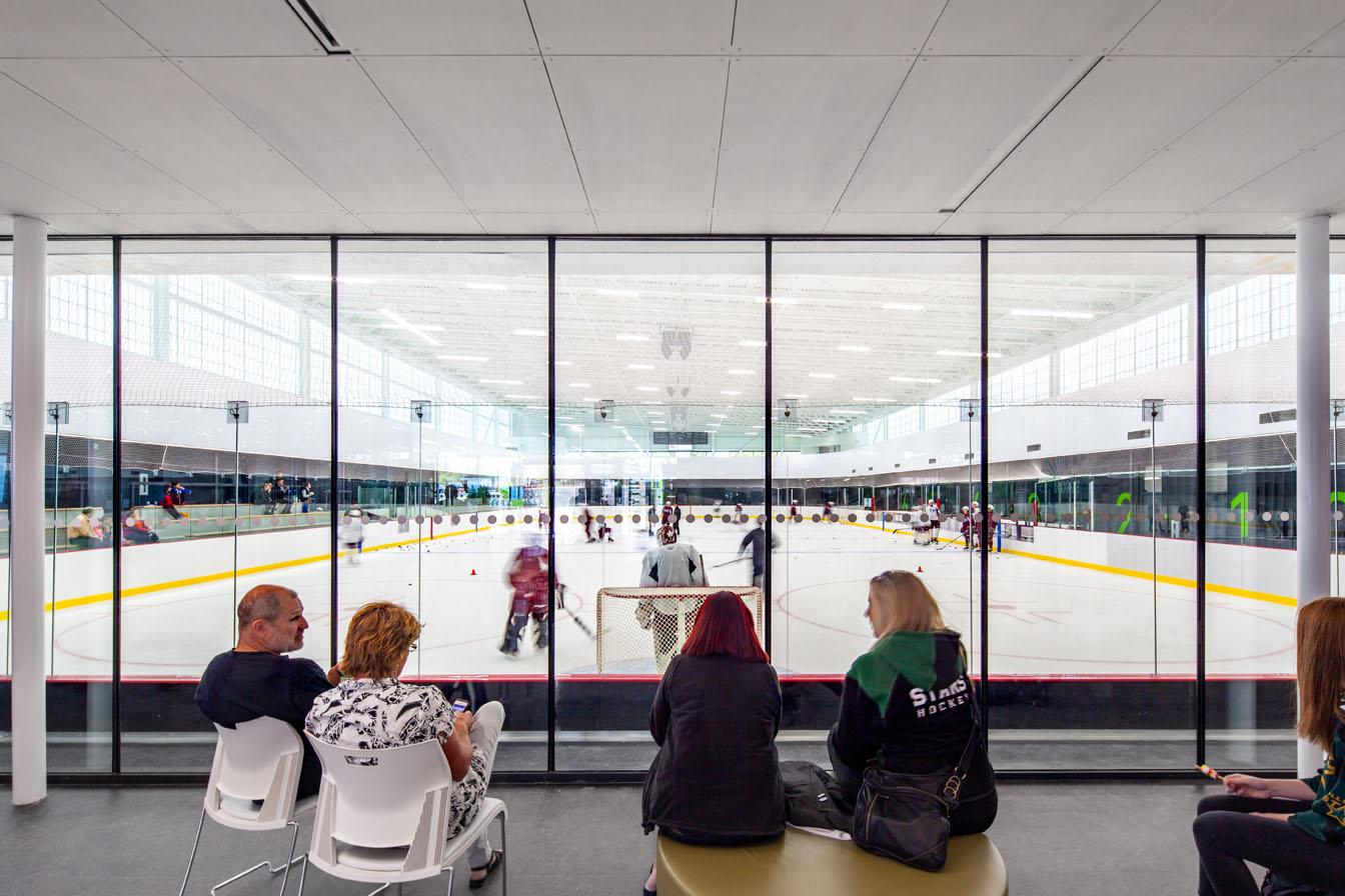 Community center interior featuring people watching hockey game through glass wall and Kalwall facade in background.