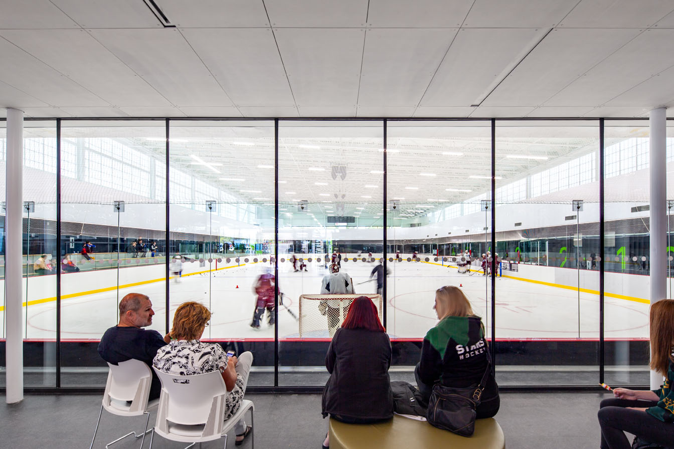Community center interior featuring people watching hockey game through glass wall and Kalwall facade in background.